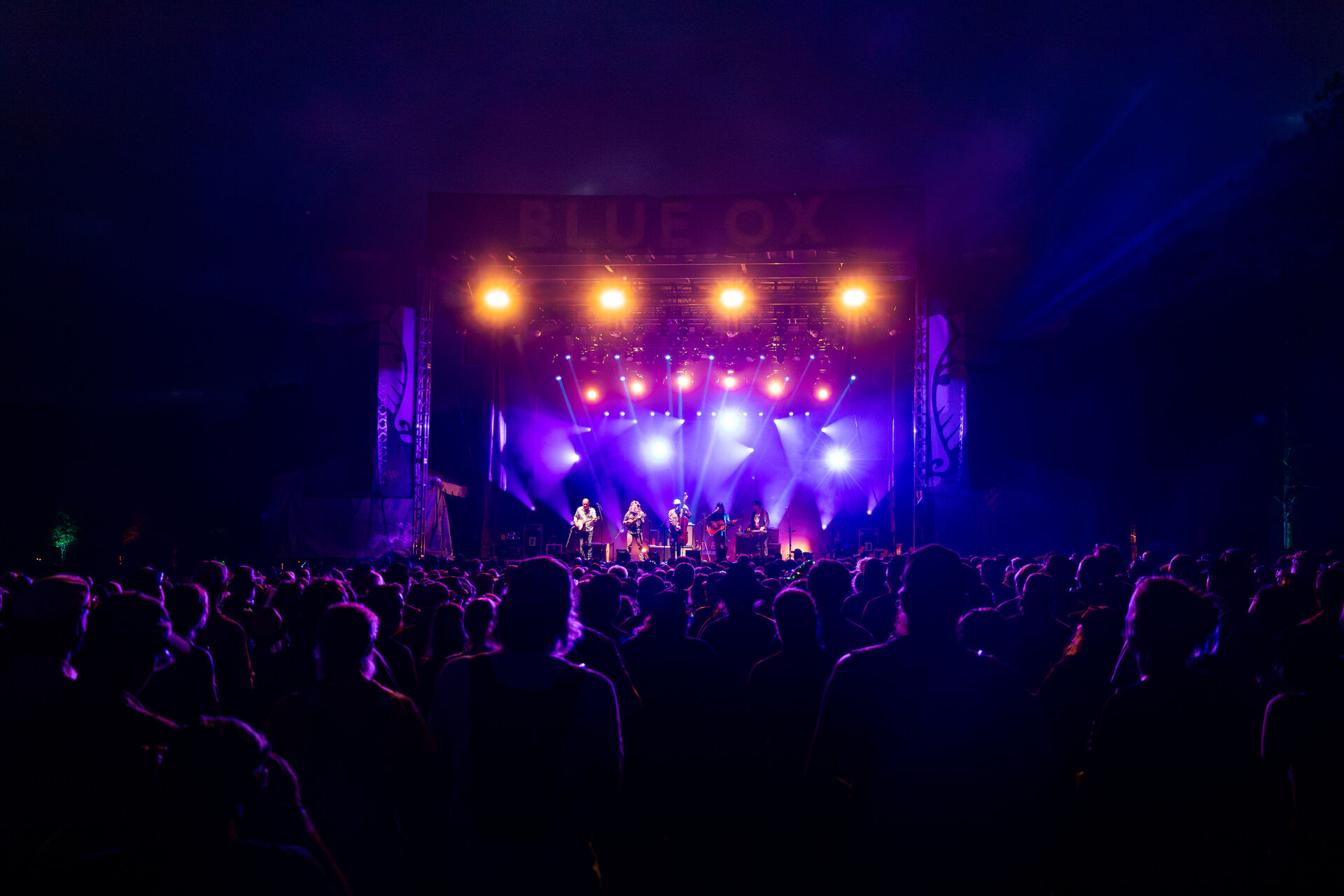 A live band on stage at the Blue Ox festival under purple and amber stage lighting, with the crowd in the foreground